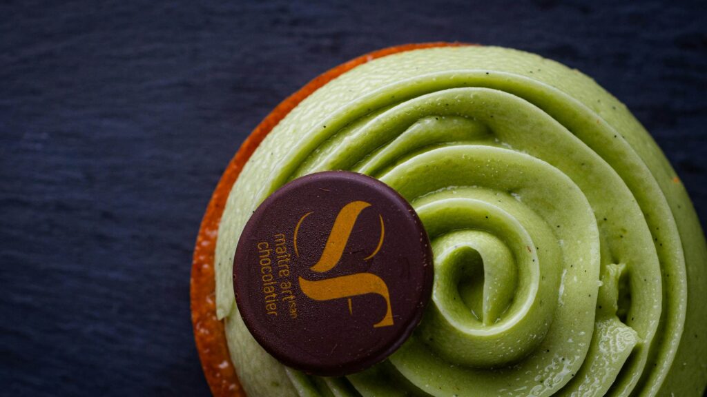 Rohelise pistaatsiakreemikattega kondiitritoode tumedal laual. Koogikesel peal logotatud šokolaadinööp.