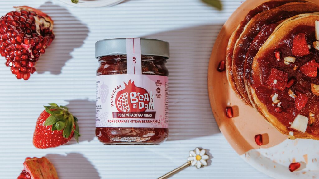 Valgel laual granaatõunamoosi purk, mille ümber taldrikul pannkoogivirn maasikatega, tükike granaatõuna, maasikas, ja lillekujuline lusika käepide.