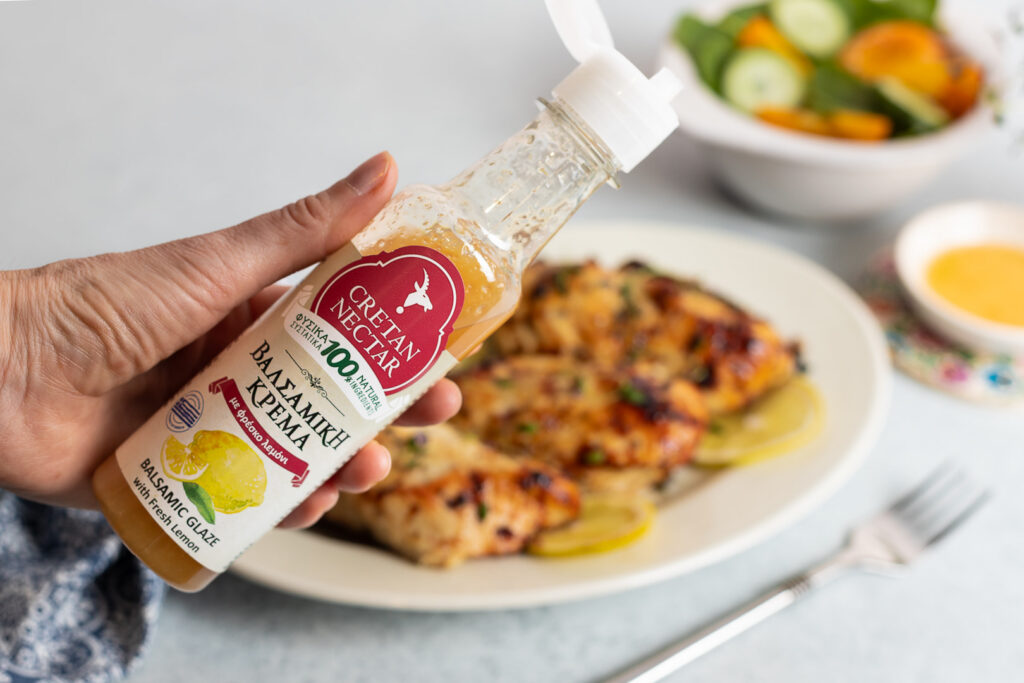 Esiplaanil suur käsi, mis hoiab Cretan Nectari sidruniga palsamikreemi pudelit. Taustal on uduselt näha valgele lauale asetatud valge taldrik marineeritud kanafileetükkidega, mille kõrvale on laotatud lõigatud sidruniviilud. Taldrikust paremal on hõbedane kahvel. Taldriku kohal paistab valges kausis värske salat.