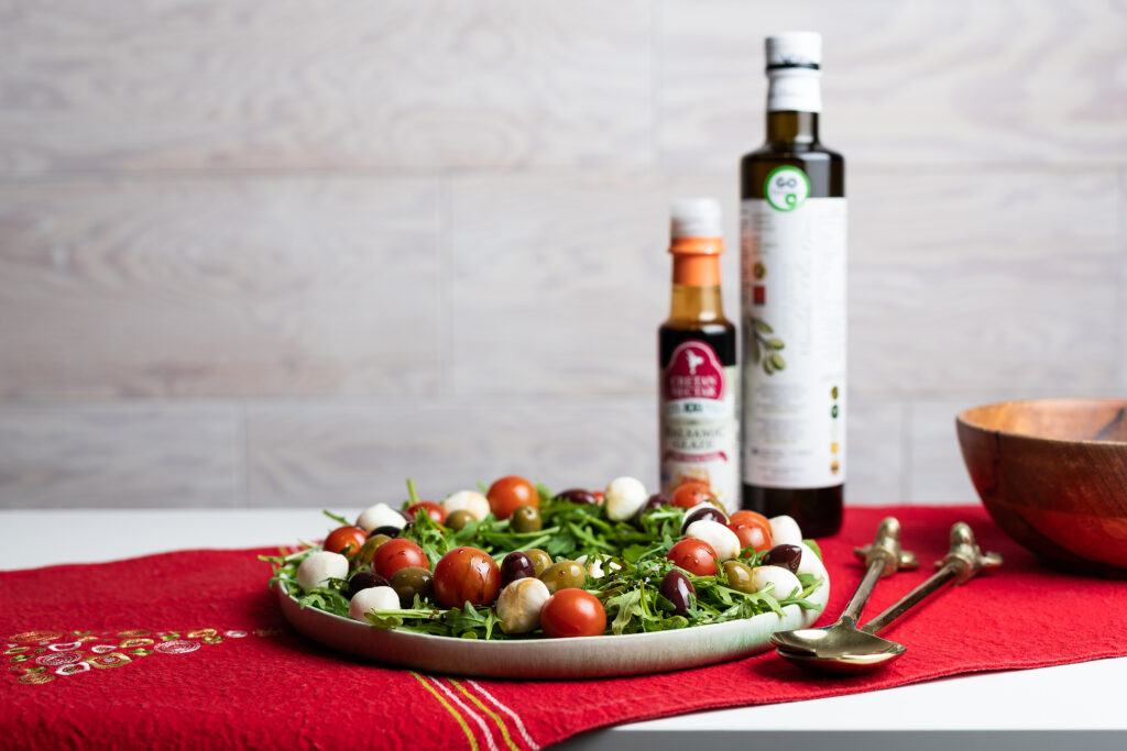 Punasel laudlinal on tomati-mozzarella salatiga taldrik, mille kõrval palsamikreemi ning oliiviõli pudel, riistad salati tõstmiseks ning kauss.