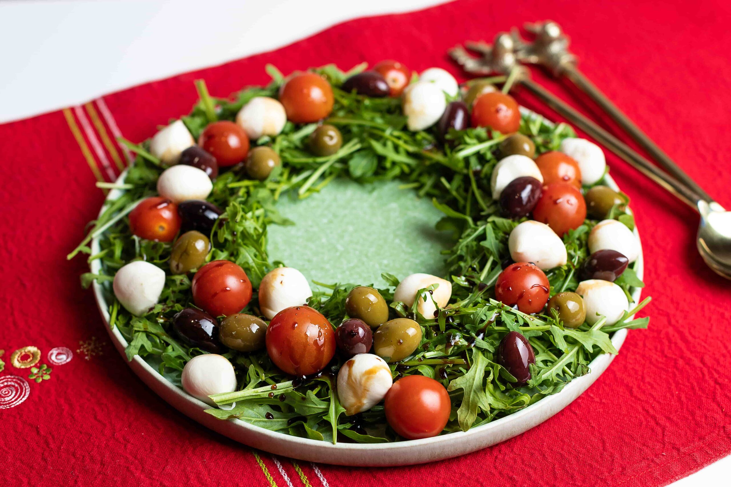 Punasel linikul on roheline taldrik, millele külgedele on laotatud rukola salatipadi. Sellele on puistatud tomatipalle, kirsstomateid ja oliive, mis on üle niristatud palsamikreemiga. Taldrikust paremal on salati tõstmiseks söögiriistad.