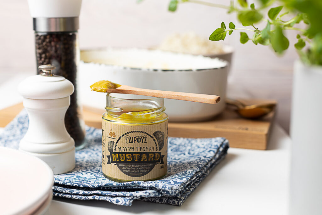 Musta trühvli maitseline sinep klaaspurgis, puidust lusikaga, taustal soola- ja pipraveski ning valge küpsetusvorm.