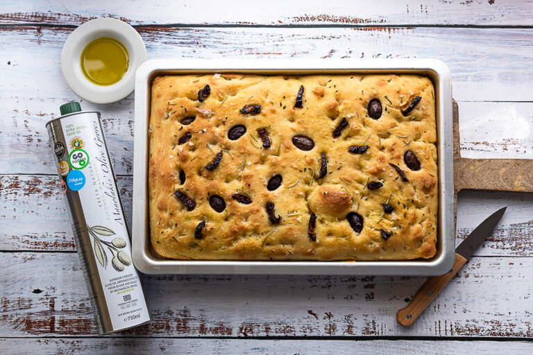 Puidust lõikelaual küpsetatud focaccia leib ahjuvormis. Millel kaunistuseks Kalamata oliivid ja rosmariin, kõrval väärisoliiviõli pudel, mille kohal kauss õliga.