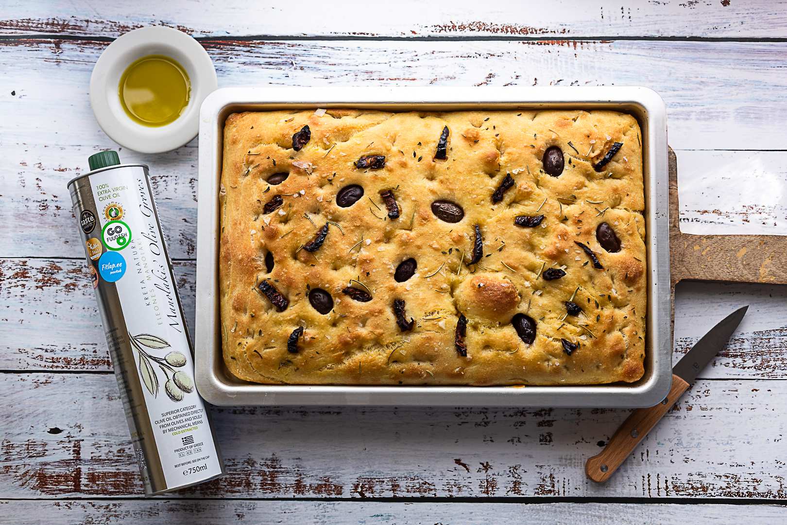 Puidust lõikelaual küpsetatud focaccia leib ahjuvormis. Millel kaunistuseks Kalamata oliivid ja rosmariin, kõrval väärisoliiviõli pudel, mille kohal kauss õliga.